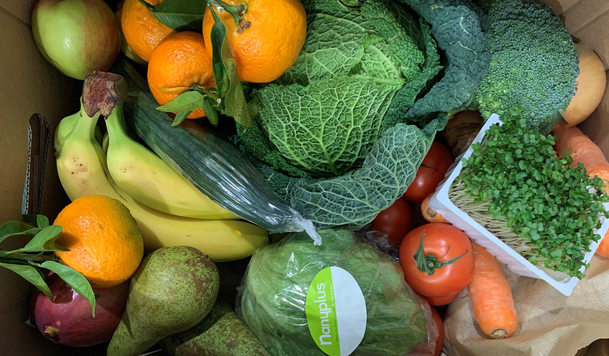 A cardboard box overflows with fresh produce, including leafy greens, vibrant oranges, ripe bananas, and a head of broccoli.