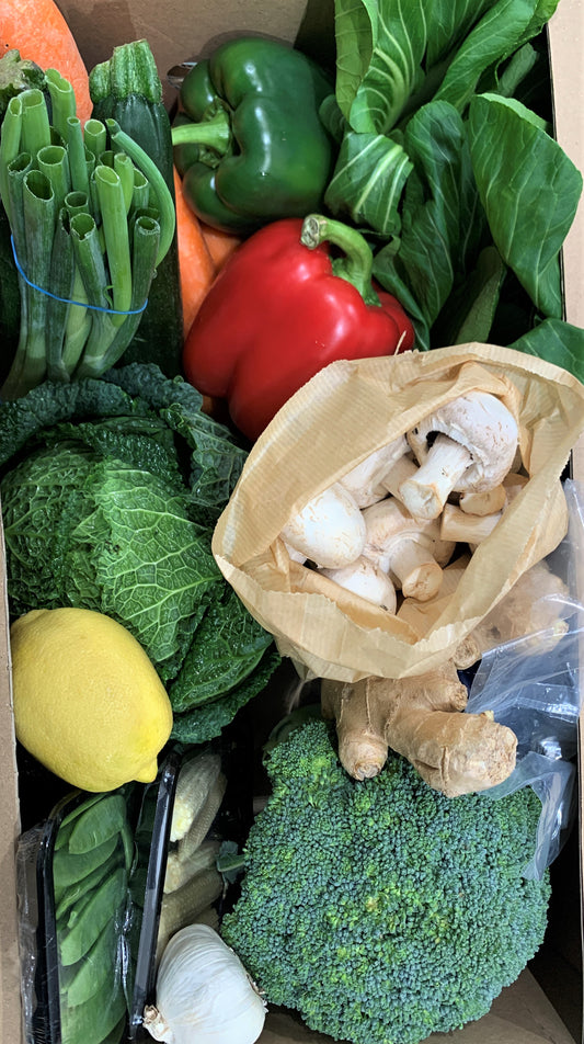 A cardboard box overflows with fresh produce, including broccoli, mushrooms, ginger, and leafy greens.